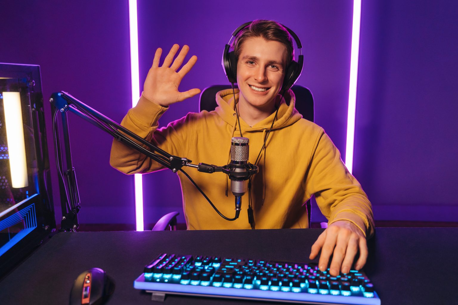Young handsome vlogger pro gamer waving hand to camera, says hello to his subscribers and followers, smiling on camera. He recording live stream of video game playthrough at home at night. Cyber sport